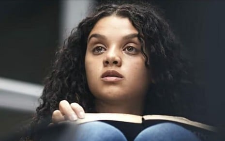 Em cena de Travessia, Danielle Olímpia está com um livro na mão, olhando para alguém com a expressão de assustada
