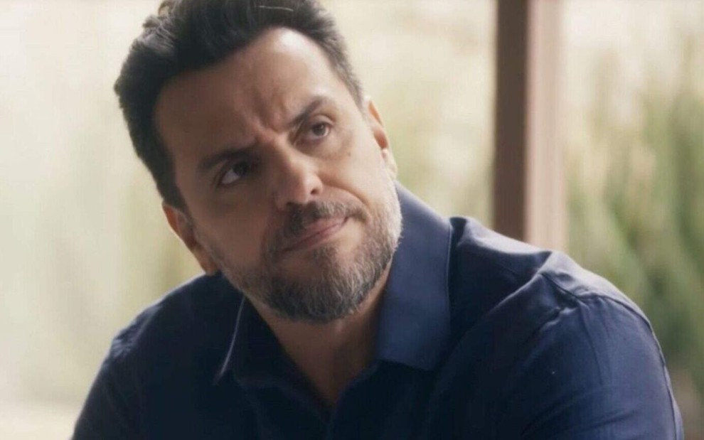 Em cena de Travessia, Rodrigo Lombardi usa blusa azul está falando com alguém com a expressão de descontentamento