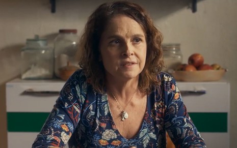A atriz Drica Moraes como Núbia em Travessia; ela está sentada a uma mesa, olhando ligeiramente para o lado com cara séria