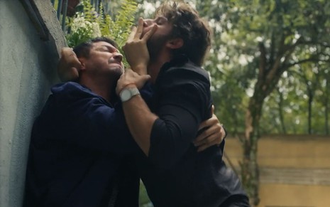 Os atores Romulo Estrela e Chay Suede como Oto e Ari em Travessia; eles estão agarrados enquanto o primeiro segura o segundo pela boca com cara de quem está fazendo força e o segundo o segura por trás