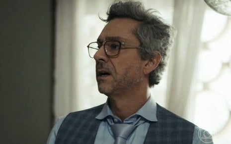 Em cena de Travessia, Alexandre Nero está com a expressão de susto; ele usa colete xadrez, blusa azul e óculos