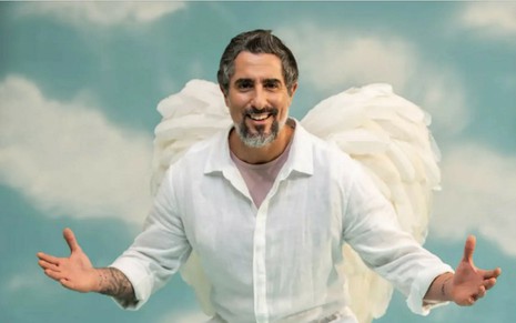 Marcos Mion com uma camisa branca e segurando uma ficha do Túnel do Amor