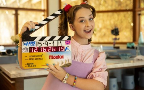 A atriz Becca Guerra com claquete na mão, cabelos presos em duas mechas, sorriso e vestido rosa, em cenário da série da Turma da Mônica