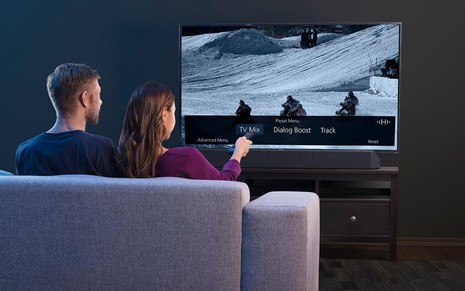 Casal no sofá ativa recursos de áudio da nova TV digital pelo controle remoto