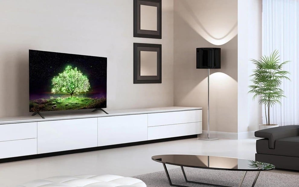 Uma sala com decoração clara em que há uma TV no rack, dois quadros na parede, uma mesa de centro e um abajur perto da janela