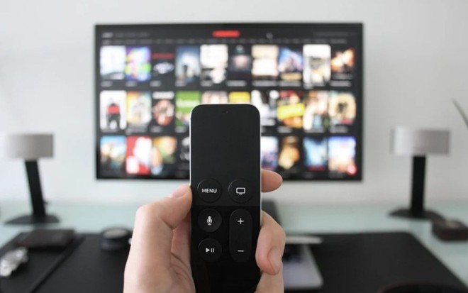 Uma mão segura um controle remoto com uma smart TV ao fundo, em uma sala bem decorada