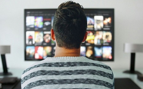 Um homem assiste TV paga com uma camisa cinza e preta e vários catálogos