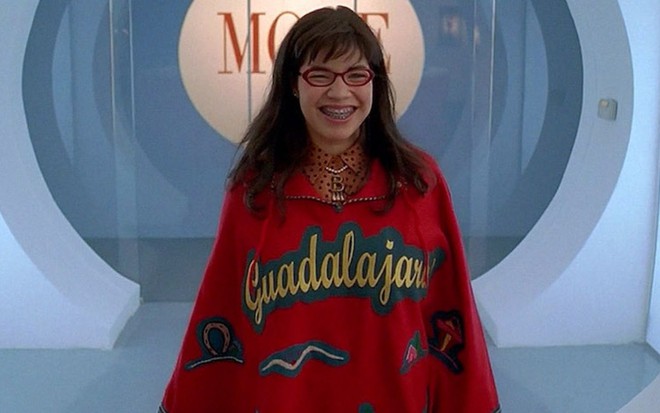 Betty Suarez (America Ferrera) usa um poncho vermelho escrito Guadalajara em amarelo; atrás dela há o logotipo da Mode em cena de Ugly Betty