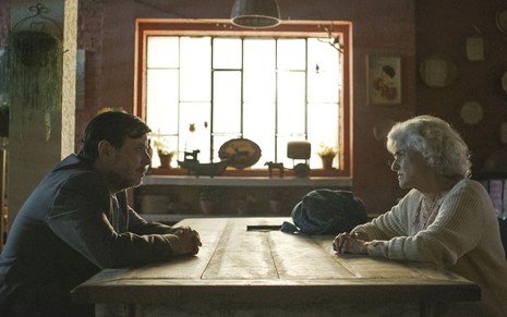 Thelmo Fernandes e Marieta Severo se encaram em cena de Um Lugar ao Sol na qual estão sentados em uma mesa em um local fechado