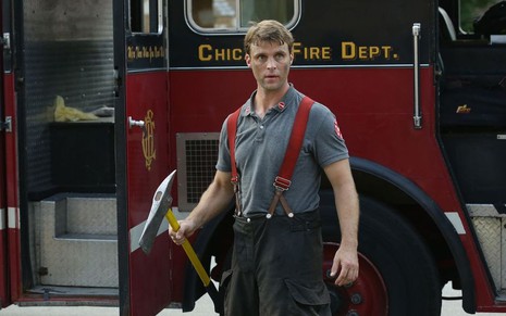 Jesse Spencer em frente a um caminhão dos bombeiros, veste uniforme e segura um machado. Ele está sujo de cinzas