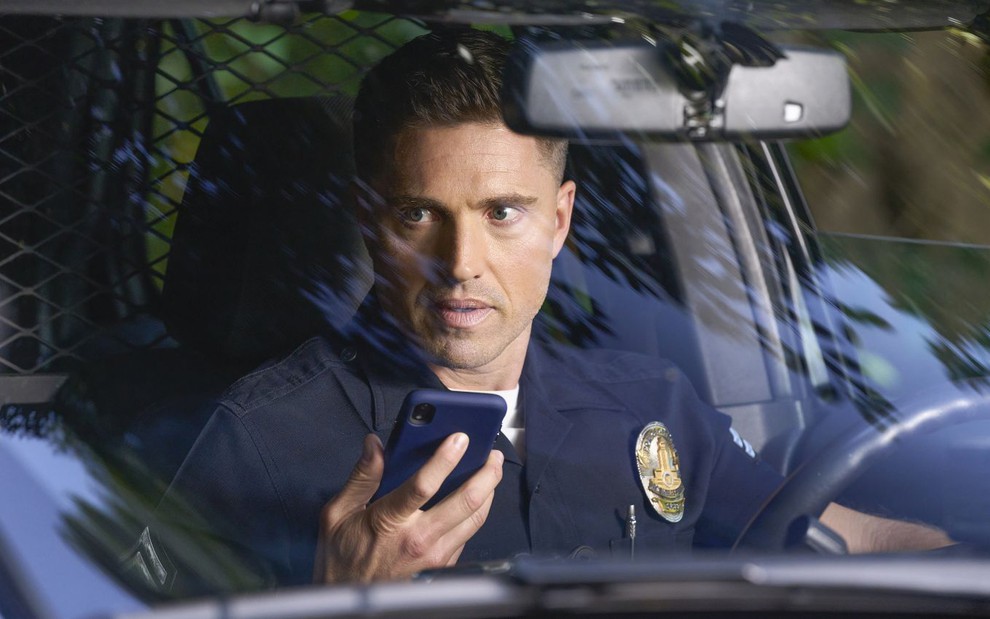 Eric Winter está dentro de uma viatura policial e segura o celular em cena de The Rookie