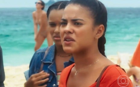 Em cena de Vai na Fé, Bella Campos está com a expressão de raiva, conversando com alguém na praia