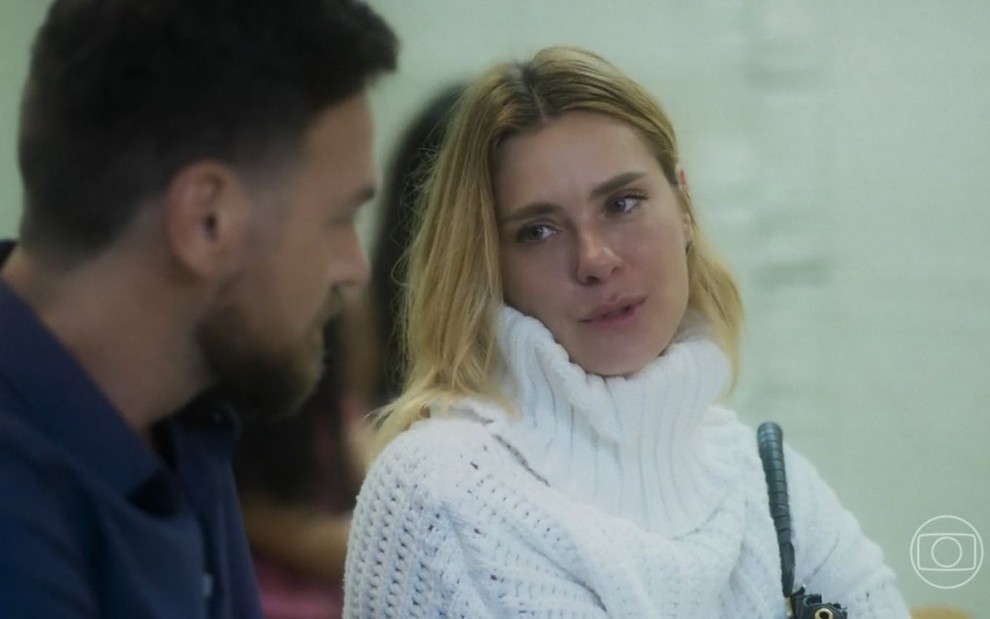 Em cena de Vai na Fé, Carolina Dieckmann usa um tricô branco de gola alta e conversa com Emilio Dantas, que usa blusa azul