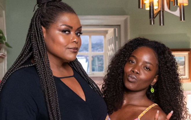 De preto, Carla Cristina Cardoso posa ao lado de Clara Moneke, que usa blusa amarela, e faz biquinho para a foto; elas estão caracterizadas como suas personagens de Vai na Fé