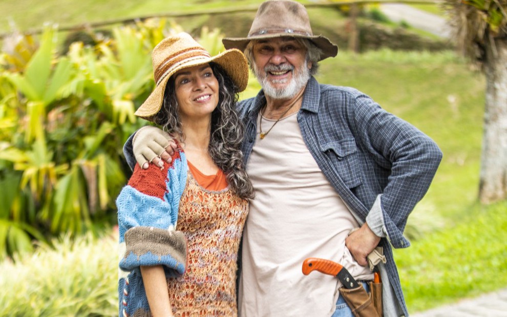 Claudia Ohana e Zécarlos Machado usam chapéu e posam no jardim de uma pousada, cenário de Vai na Fé