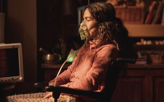 Dora (Claudia Ohana) em cena de Vai na Fé, com aparelho para respirar no sorsto, expressão séria, sentada, de perfil