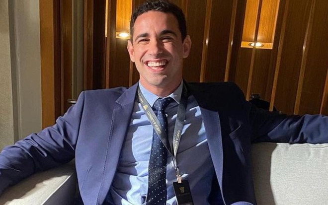 O ator Giuliano Laffayette sorrindo em estúdio de Vai na Fé, de terno e gravata, sentado em sofá