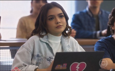 Com blusa de gola branca e jaqueta jeans, Mel Maia está em sala de aula com notebook aberto em cena de Vai na Fé