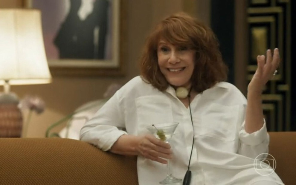 De blusa branca e segurando um martini, Renata Sorrah está em cena de Vai na Fé, sentada em um sofá marrom