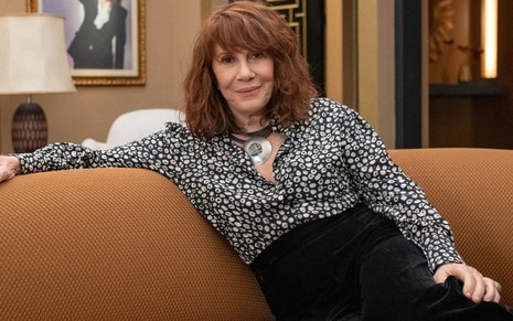 Com blusa estampada branca e preta e calça preta, Renata Sorrah está sentada em uma sofá marrom no cenário de Vai na Fé