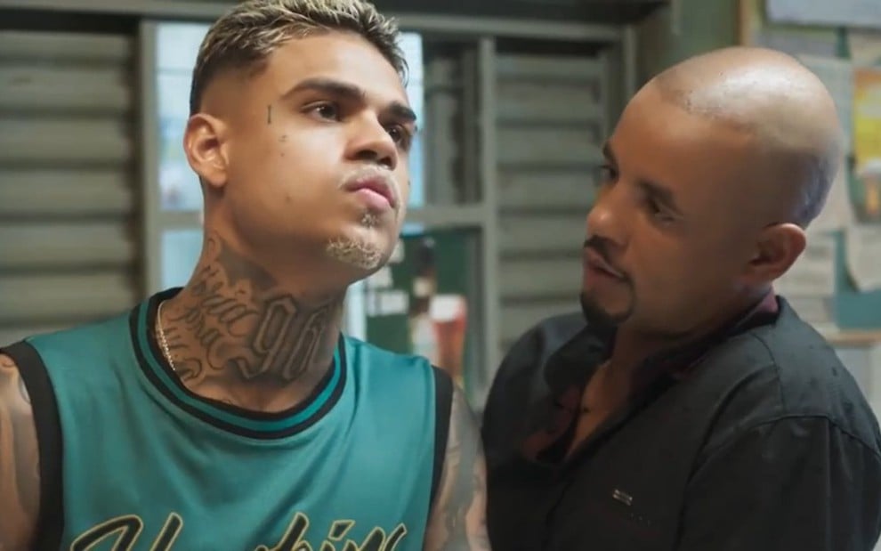 Hugo (MC Cabelinho) e Orfeu (Jonathan Haagensen) lado a lado, Cabellinho com olhar vago, Haagensen olhando sério para o colega, em cena da novela Vai na Fé