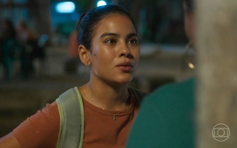 Em cena de Vai na Fé, Bella Campos está com uma mochila nas costas, blusa marrom e cabelo preso; ela fala com alguém de costas