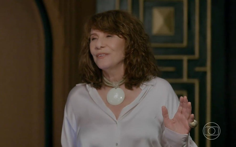 Em cena de Vai na Fé, Renata Sorrah usa blusa branca e está falando com alguém