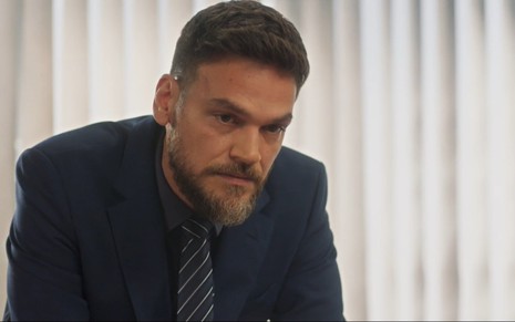 O ator Emilio Dantas com expressão séria em cena de Vai na Fé, de terno e gravata em escritório