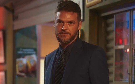 O ator Emilio Dantas com expressão séria em cena de Vai na Fé, de terno e gravata