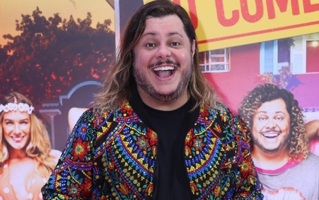 Marcos Majella com um cabelo longo e rindo durante gravação de Vai Que Cola