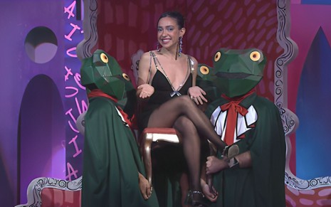 Valentina Bandeira sorrindo, de vestido preto curto, sentada numa cadeira carregada por dois homens com fantasia de sapo, no estúdio do programa Beija Sapo