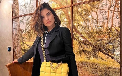 A atriz Vanessa Giácomo, que estará em Travessia, posa na frente de uma janela usando uma bolsa grande e amarela