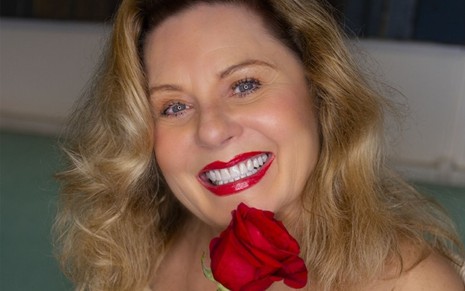 Atriz Vera Fischer veste blusa branca, sorri com batom vermelho e segura uma rosa vermelha em mãos