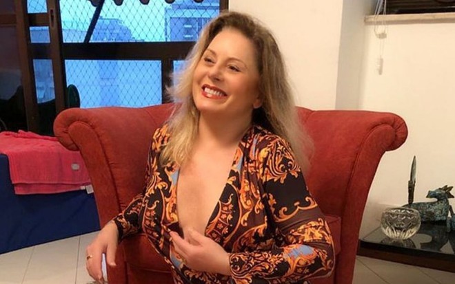 Vera Fischer, vestida de um body estampado e decotado, sorri encostada em um sofá vermelho
