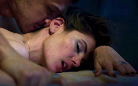 A atriz Camila Queiroz está deitada numa cama, com os olhos fechados, enquanto Romulo Estrela beija seu pescoço em gravação de Verdades Secretas 2
