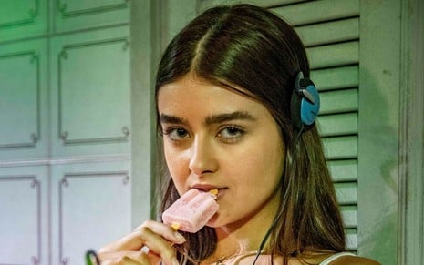 Júlia Byrro em cena de Verdades Secretas 2: caracterizada como Lara, atriz está com regata branca e picolé rosa na boca
