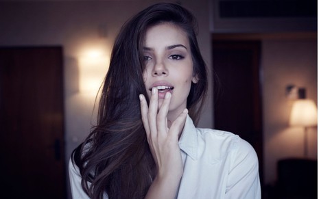 De camisa branca de botões, Camila Queiroz com cabelo para o lado direito e põe a mão na boca com os lábios entreabertos