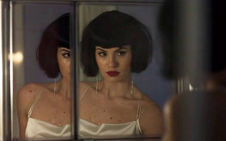 Atriz Camila Queiroz usa peruca preta e batom vermelho enquanto se olha no espelho em cena de Verdades Secretas 2