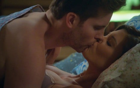 Deitados na cama, Alan (Thiago Fragoso) beija Kyra/Cleyde (Vitória Strada) em Salve-se Quem Puder