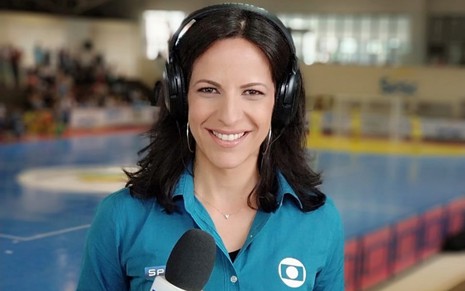 Viviane Costa com uma camisa azul, segurando o microfone da Globo em uma arena esportiva