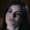 Camila Queiroz como Angel. Ela usa um vestido preto, um cabelo longo e olha de forma sedutora para a câmera.