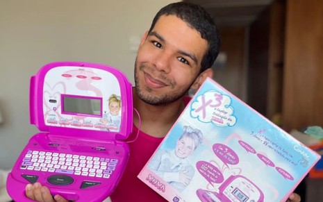 Vinicius Fernandes está sentado em uma cama e segura com as duas mão um laptop rosa e a embalagem do brinquedo