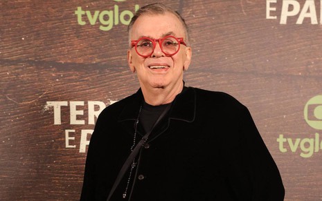 O autor Walcyr Carrasco posa para foto no evento de lançamento da novela Terra e Paixão, com terno e camisa pretos, óculos vermelhos, sorrindo