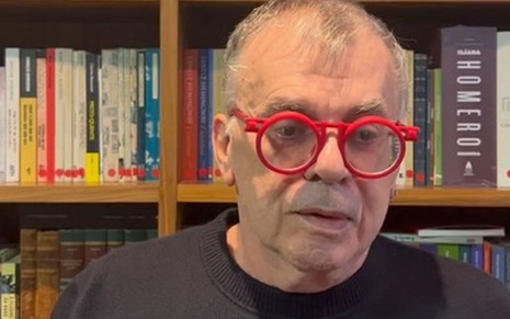 Com óculos vermelho, Walcyr Carrasco fala na frente de uma estante cheia de livros