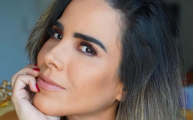 Wanessa Camargo está com uma das mãos apoiadas no queixo e sorri discretamente