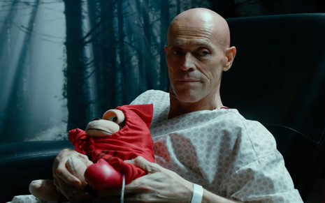 Willem Dafoe careca, com roupa de hospital, sentado, segurando um bichinho de pelúcia vermelho