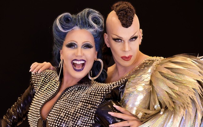 Xuxa Meneghel e Ikaro Kadoshi posam um ao lado do outro. Ambos usam peruca e Xuxa está com uma jaqueta preta cheia de brilhantes. Ikaro veste um vestido dourado com ombreiras que simulam penas brilhantes.