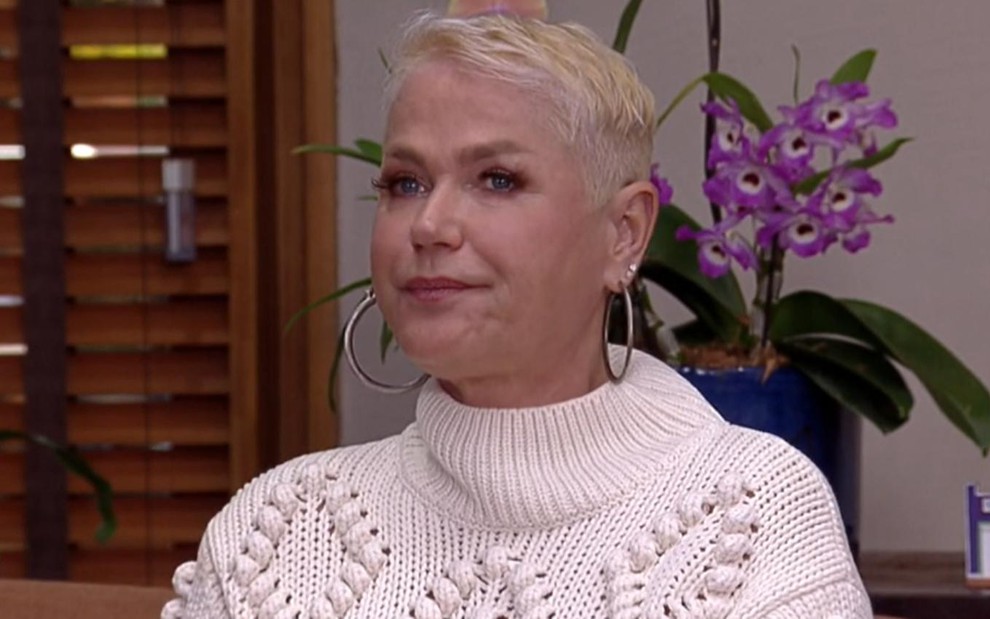 A apresentadora Xuxa com expressão séria, clusa de lã branca, no estúdio do É de Casa
