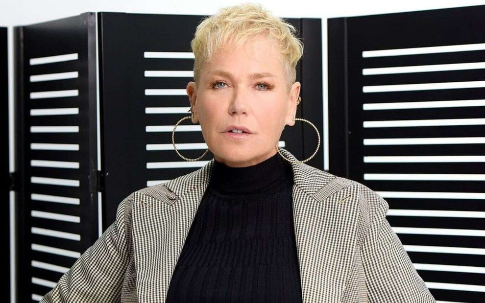 Imagem com a apresentadora Xuxa vestindo blusa preta e blazer xadrez nas cores bege e preto