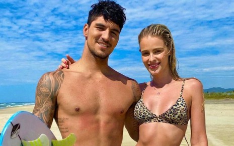 Gabriel Medina ao lado de Yasmin Brunet numa praia, eles estão abraçados e sorrindo; ele sem camisa e segurando uma prancha, ela de biquíni de oncinha